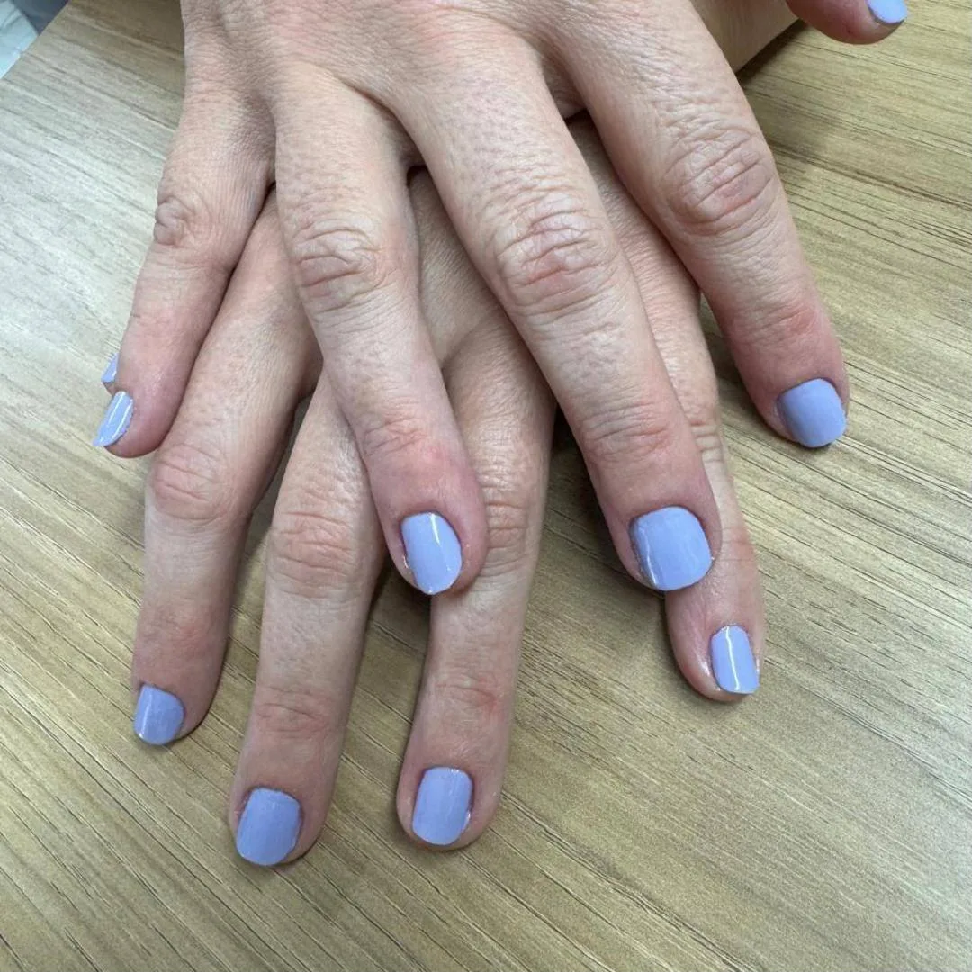 Mãos sobrepostas com unhas curtas em lilás claro, sobre uma mesa de madeira